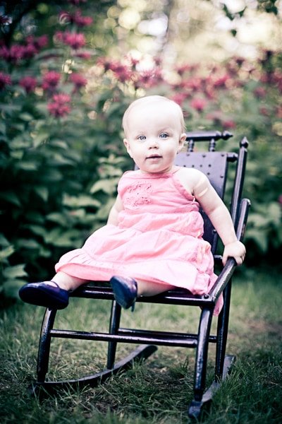 A baby in a pink dress sits on a small wooden rocking chair outdoors. The background has green foliage and red flowers, creating a serene garden setting.