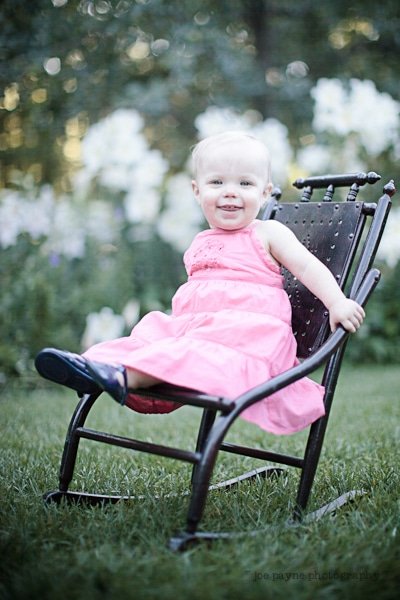 A smiling toddler wearing a pink dress sits on a wooden chair in a garden. The background features lush greenery and blooming white flowers.