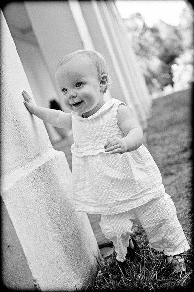 Black and white photo of a smiling baby standing outdoors, wearing a light dress and pants, leaning against a column. The baby is looking to the side, with one hand resting on the column and grass visible in the background.