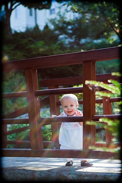 A smiling toddler in a white outfit stands on a wooden bridge, holding onto the railing. Sunlight filters through the trees, casting colorful lens flares across the scene. The background is lush and green.