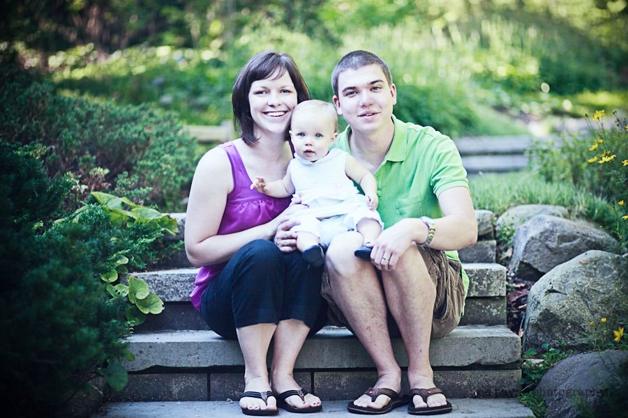 A woman, man, and baby sit together on outdoor steps surrounded by greenery. The woman in a purple top holds the baby, while the man in a green shirt sits beside them. They all smile at the camera.