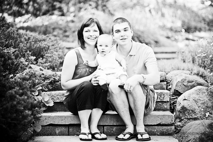 A black and white photo of a smiling family sitting on stone steps outdoors. The mother holds a baby in the center, and the father sits beside them. Lush greenery surrounds the steps.