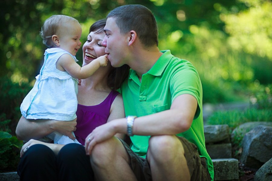 A cheerful family sits outdoors on a sunny day. A smiling woman holds a baby dressed in white, reaching for the mans nose. The man, wearing a green shirt, smiles and gently leans toward the baby. Green foliage surrounds them.