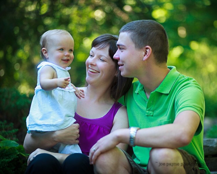 A smiling family with a young child outdoors. A woman in a purple top holds a baby in a white dress, while a man in a green shirt leans in, all appearing joyful and relaxed against a lush green background.