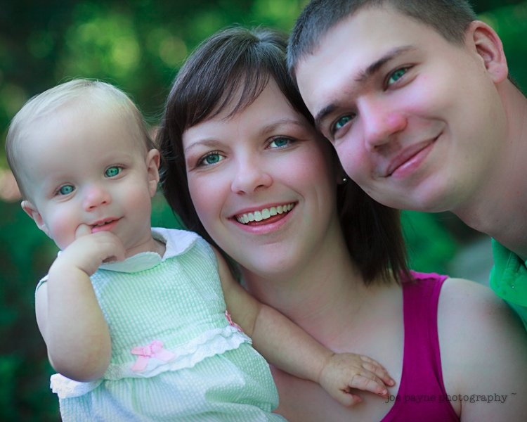 A woman in a pink top and a man in a green shirt pose joyfully with a smiling baby in a light green dress. They are outdoors with a blurred green background.