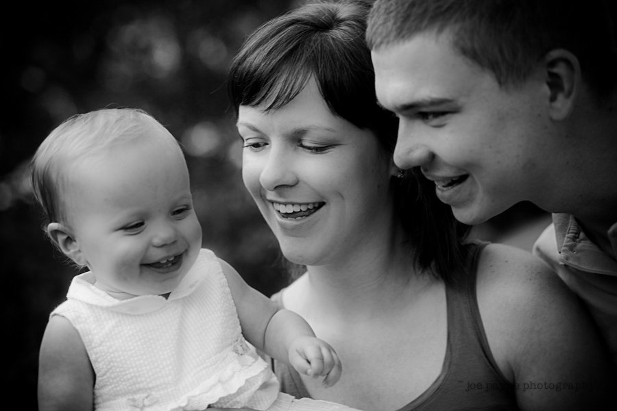 A joyful black and white image of a smiling family: a woman, a man, and a baby. The baby, in a light dress, is looking at the woman, who appears to be the mother. The man, likely the father, is on the right, looking fondly at the baby.