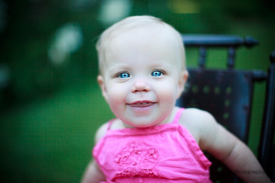 A smiling baby with bright blue eyes and wearing a pink outfit sits on a chair outdoors against a blurred green background.