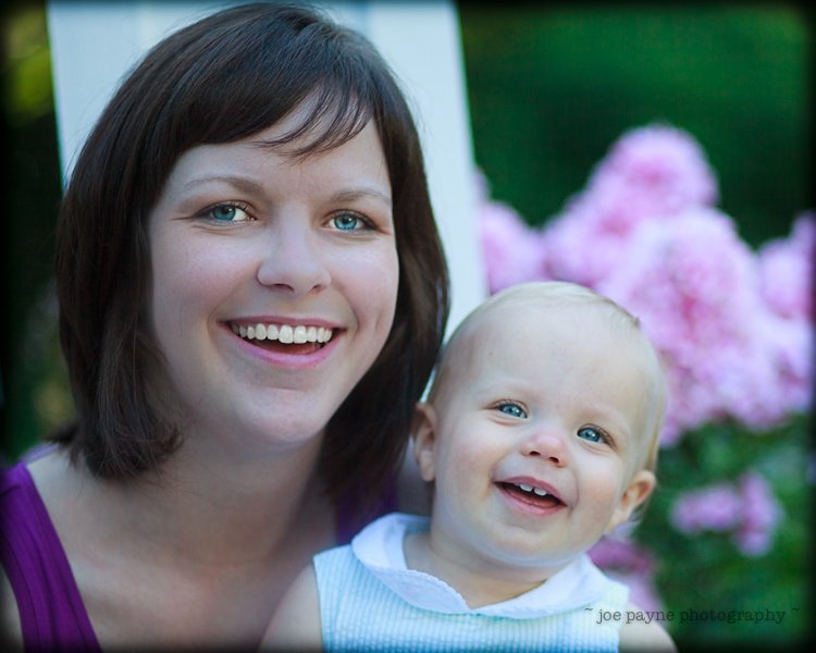 A woman and a baby smiling outdoors in front of blooming pink flowers. The woman has short brown hair and is wearing a purple top, while the baby, wearing a light-colored outfit, has light hair and is smiling brightly.