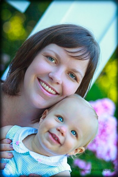 A smiling woman holds a happy baby in an outdoor setting. The baby has bright blue eyes and is wearing a sleeveless white outfit. The background features green foliage and pink flowers.