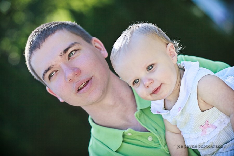 A man in a green shirt holds a smiling baby wearing a white dress with pink accents. They are outdoors with a blurred green background.