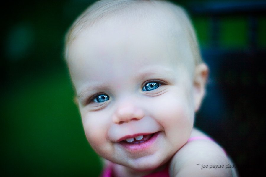 A smiling baby with bright blue eyes looks directly at the camera. The background is blurred with a green hue, highlighting the baby’s joyful expression.