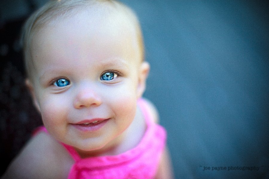 A close-up of a smiling baby with blue eyes, wearing a pink outfit. The background is blurred, focusing on the childs joyful expression.