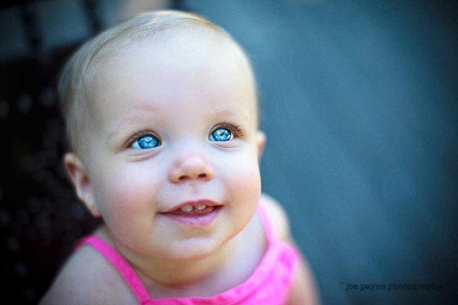 A baby with bright blue eyes and a pink outfit looks up with a joyful expression, set against a softly blurred background.