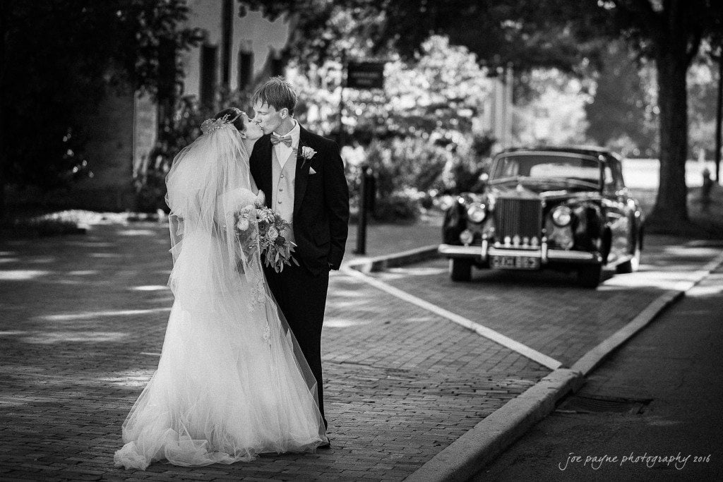 A bride and groom share a kiss on a cobblestone street, with the groom in a black suit and the bride in a flowing white gown and veil. A vintage car is parked nearby under a tree. The scene is in black and white.