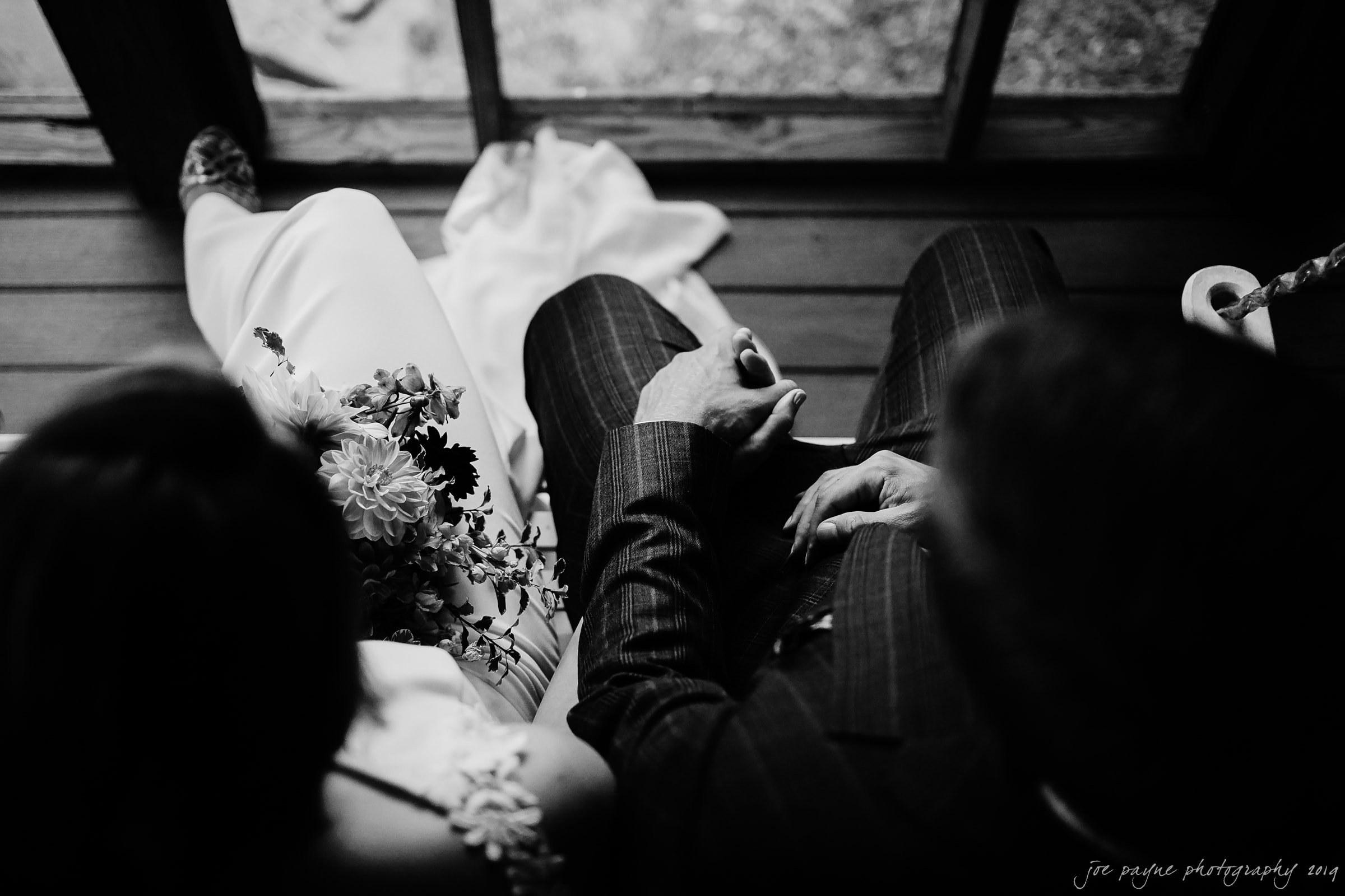 A black and white image of a couple sitting closely, presumably during a wedding. The person on the left holds a bouquet, while the person on the right wears a suit. Theyre seated on a wooden floor near large windows.