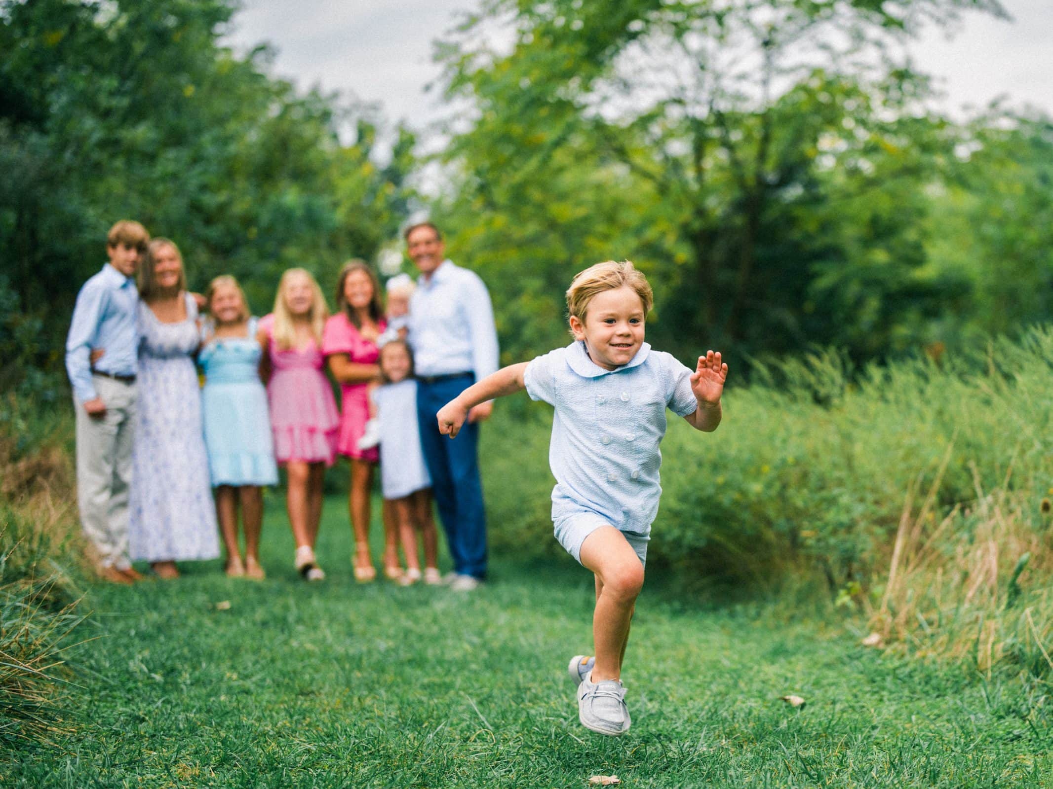 A young child in a light blue outfit runs energetically towards the camera on a grassy path. In the background, an out-of-focus group of people stands together smiling, surrounded by greenery. The scene is bright and cheerful.