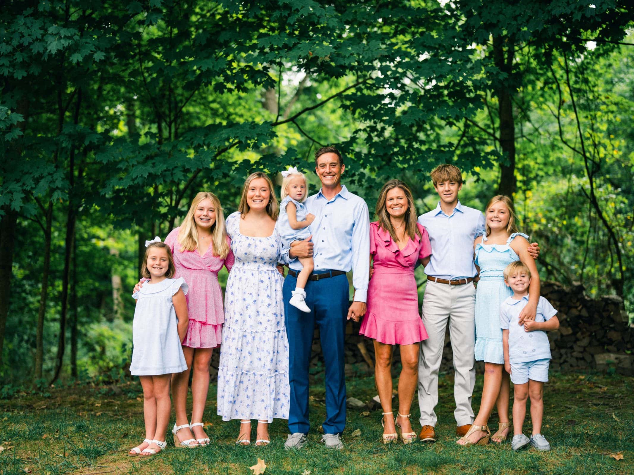 A family of eight poses outdoors in a forest setting. The group includes four adults and four children. Some wear summer dresses, and others are in shirts and pants. They are all smiling, standing on a grassy area with trees in the background.