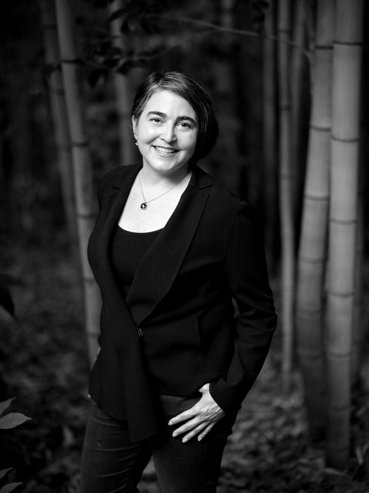 A woman wearing a dark blazer and top stands smiling in a forested area with tall bamboo trees in the background. The image is in black and white, creating a serene and natural atmosphere.