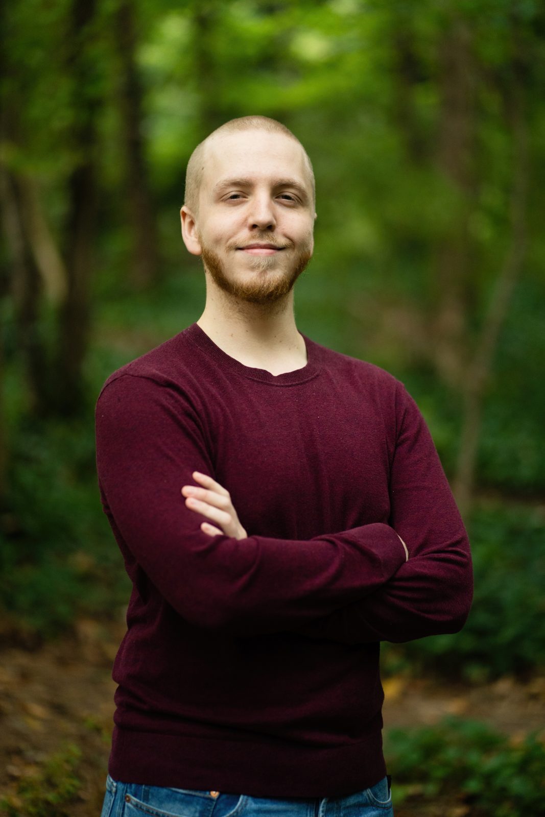 A person with a shaved head and beard stands outdoors in a forest, wearing a maroon sweater and blue jeans. Their arms are crossed, and they are smiling slightly. The background is lush and green, suggesting a wooded area.