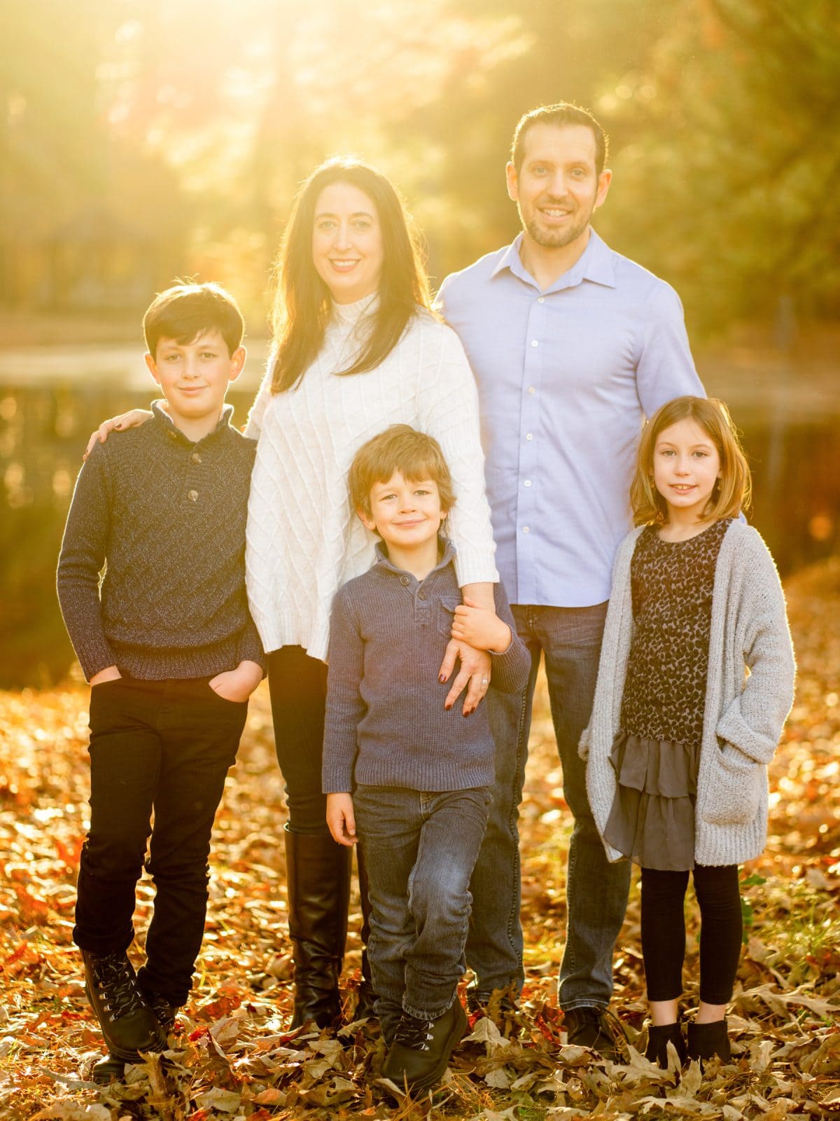 A family of five stands outdoors on a sunny autumn day, surrounded by fallen leaves. The parents stand behind their three children, who are smiling and wearing cozy sweaters. Trees and a reflective lake are in the background.