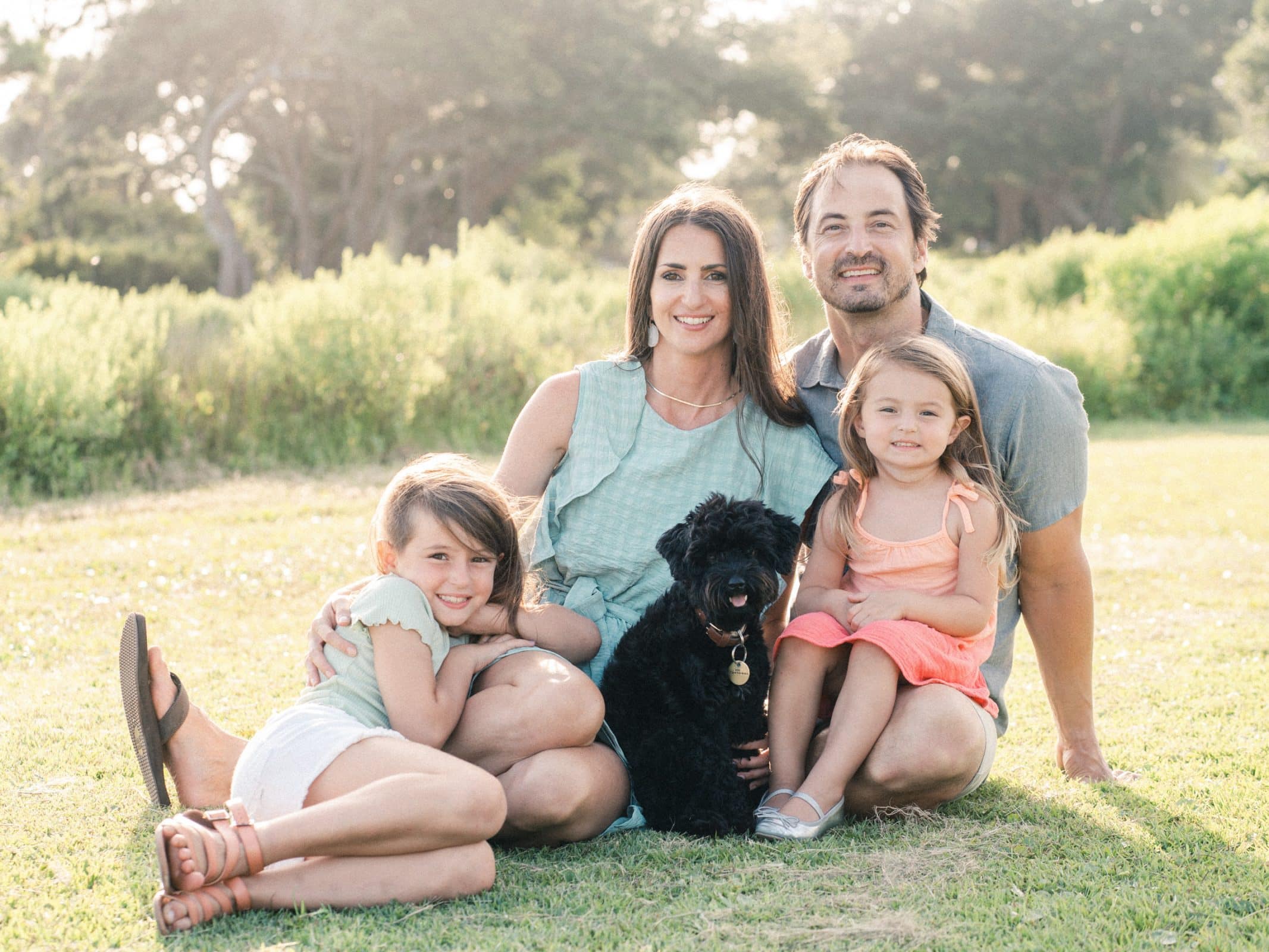 A family of four, with two young children and a black dog, sits together on a grassy field. The adults are smiling and the children look content. The sunlit background features lush greenery and tall trees.