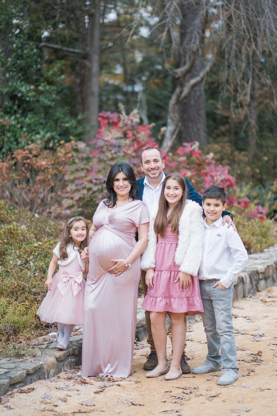 A family poses outdoors on a path surrounded by trees and foliage. The mother, in a pink dress, is pregnant. A father, a young girl, a teenage girl, and a boy smile beside her, all dressed in coordinated pink and white outfits.