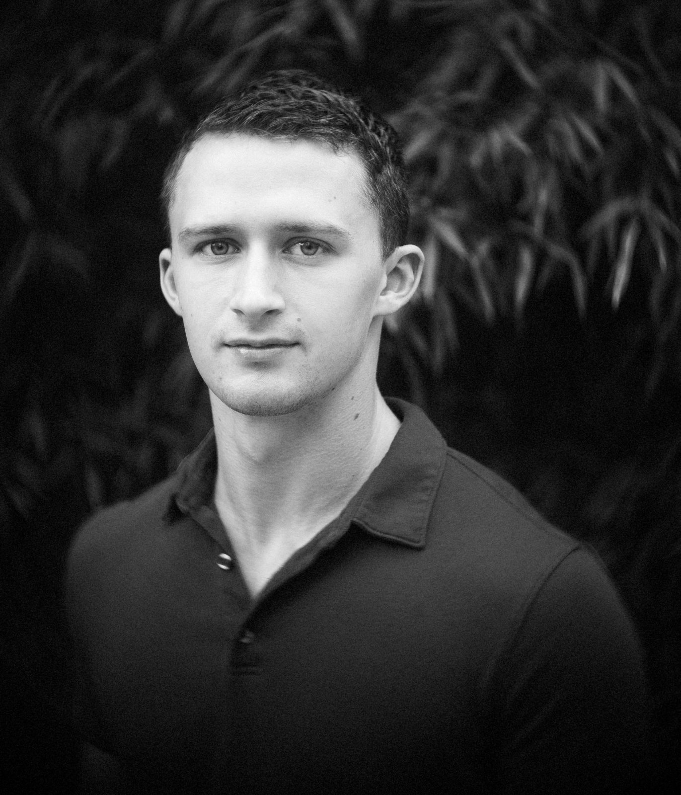 A black-and-white portrait of a person with short hair wearing a dark collared shirt, captured in the classic style of Raleigh headshot photography. The softly blurred foliage background enhances their neutral expression as they look directly at the camera.