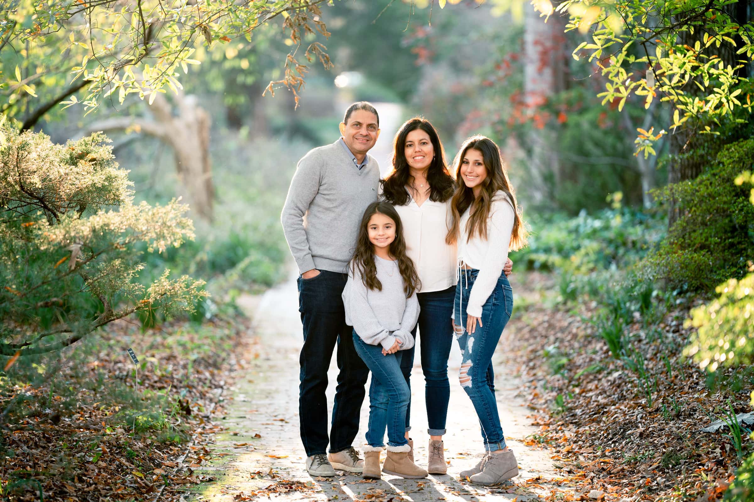 A family of four poses on a forest path near Raleigh, with two adults and two children in casual clothing. Surrounded by greenery and autumn foliage, sunlight filters through the trees, creating a perfect setting reminiscent of professional photography ideal for capturing cherished headshots.