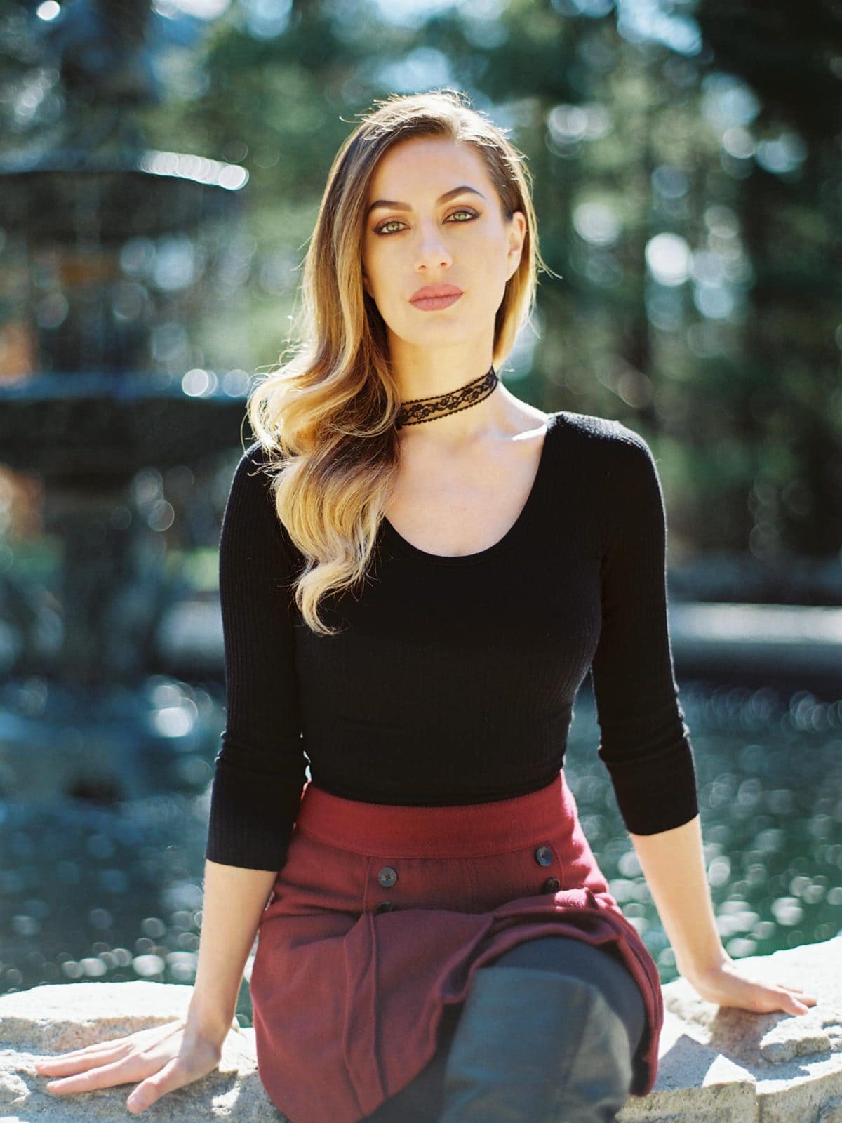 A woman with long, wavy hair sits outdoors on a stone ledge. Her black top, burgundy skirt, and choker necklace add elegance against the blurred backdrop of trees and a fountain. This picturesque scene could be perfect for headshot photography in the Raleigh Durham area.