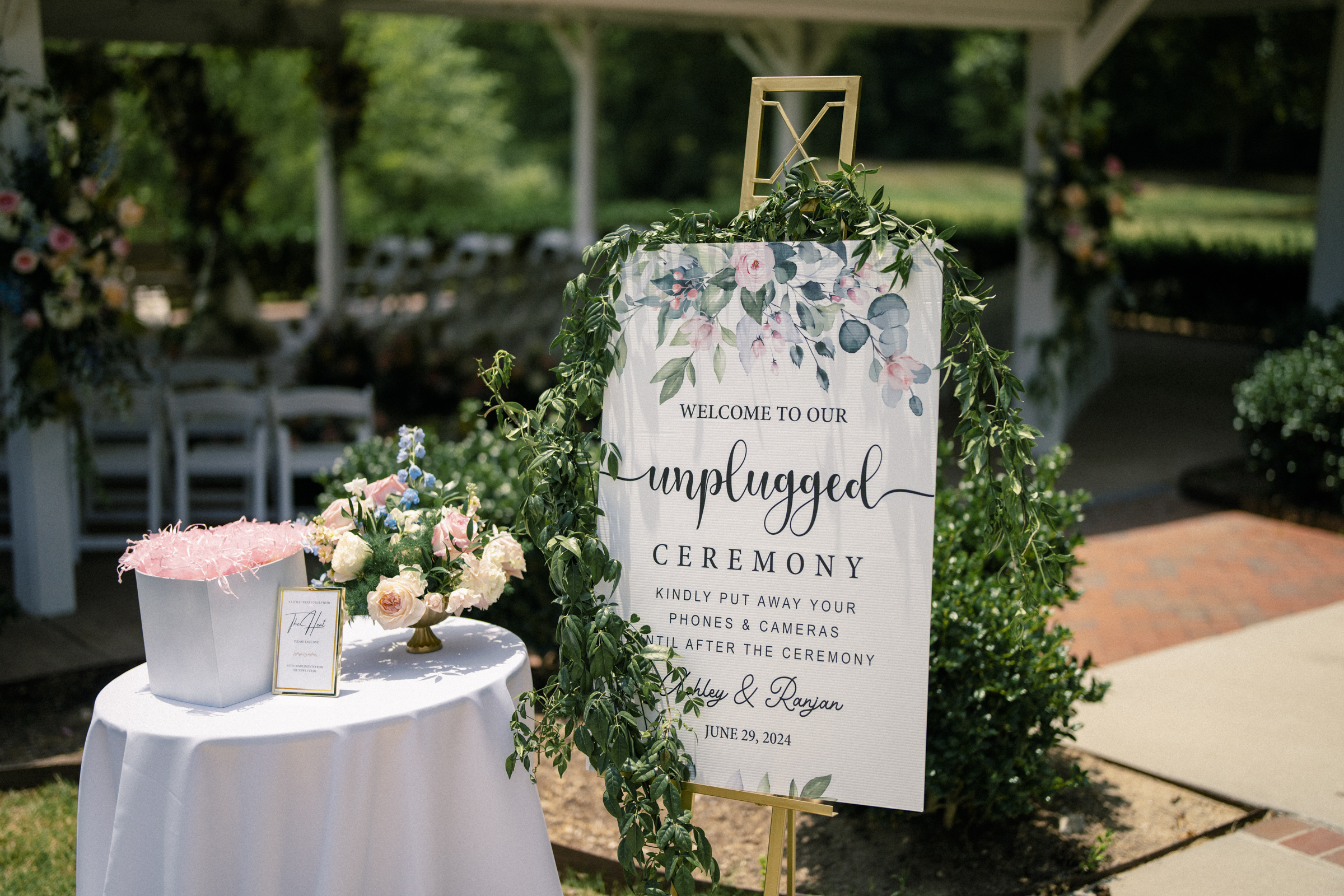 An outdoor wedding display showcases a sign reading Welcome to our unplugged wedding ceremony, adorned with elegant floral decorations. Nearby, a table holds a box, guestbook, and flower arrangement. Chairs and lush greenery decorate the background under a charming gazebo.