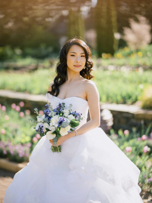 A woman in a white wedding dress holds a bouquet of purple and white flowers during her bridal session. She stands outdoors in a garden with blurred greenery and blooming flowers in the background, looking directly at the camera. The scene is bathed in soft, warm light.