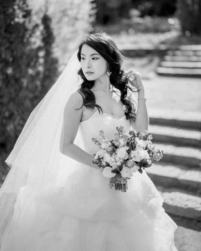 A black-and-white photo from a bridal session captures a bride in a flowing wedding gown, holding a bouquet. With long, wavy hair and a veil, she gazes serenely to the side. Stone steps and greenery blur softly in the background.