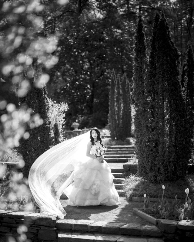 A bride in a flowing, layered wedding gown stands on a garden pathway, holding a bouquet. Her long veil catches the wind, trailing elegantly behind her. Tall trees and lush greenery frame the scene, creating a serene atmosphere perfect for enchanting bridal sessions.