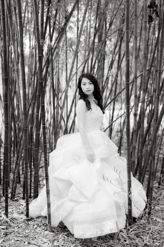 In a serene bridal session, a woman in a large, layered white gown stands among tall bamboo stalks. The black and white image adds a classic, timeless feel. Her hair cascades gracefully over her shoulders as she exudes tranquility amid the forest's quiet embrace.