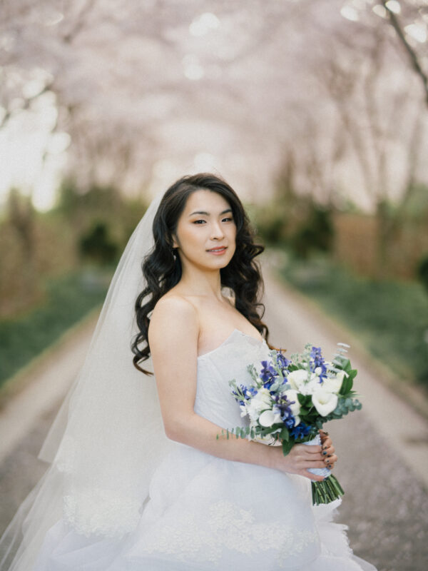 During the bridal sessions, a bride in a strapless wedding dress holds a bouquet of white and purple flowers. Her long, dark hair cascades over her shoulders as she stands on a tree-lined path with pink blossoms, creating a serene and romantic atmosphere.