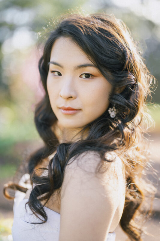 A woman with long, wavy dark hair and a subtle smile stands outdoors in soft lighting, reminiscent of intimate bridal sessions. With blurred greenery in the background, she wears a sleeveless top and delicate earrings, exuding a calm and serene expression.