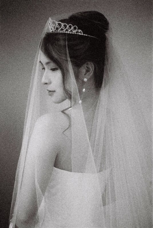 A black and white bridal session portrait of a bride in a strapless dress, captured from the side. She wears a tiara and a long veil, with her hair elegantly styled up. The image has a soft, grainy texture, adding a vintage feel.