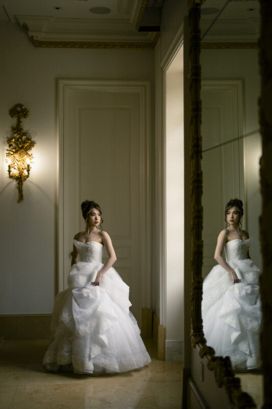 A bride, during her bridal session, stands near a mirrored wall in an elegant room, wearing a white, strapless wedding gown with a voluminous skirt. Soft lighting casts a warm glow, highlighting her reflection in the mirror and the ornate wall sconce beside her.