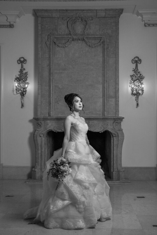 In a refined black-and-white scene, a bride stands in her elegant lace wedding gown, bouquet in hand, gracing the timeless charm of an ornate fireplace. This sophisticated setting is perfect for capturing exquisite bridal sessions with its classic ambiance and decorative wall sconces.