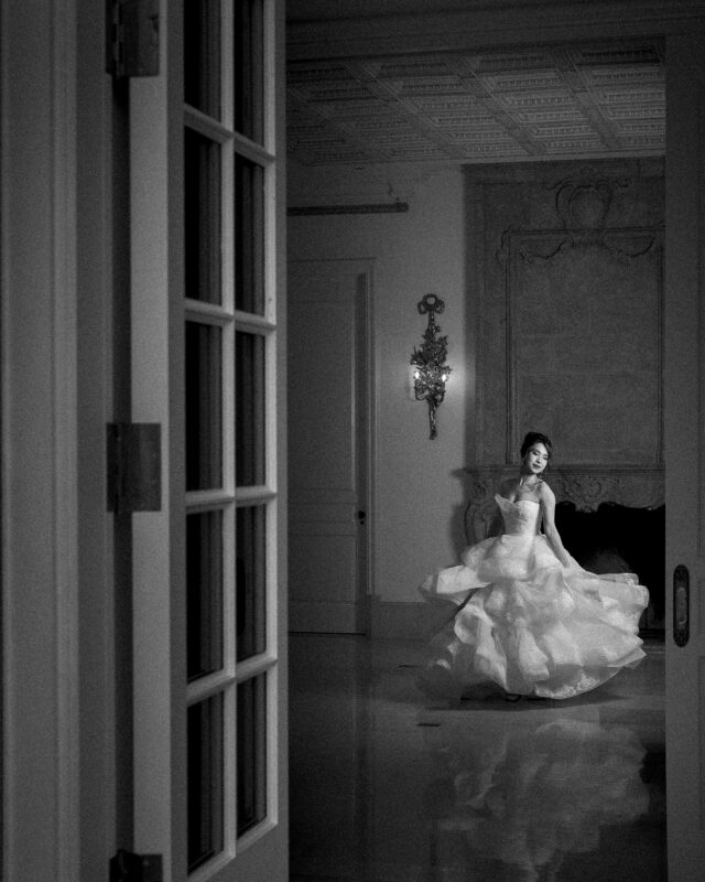 A black and white photograph captures a woman in a flowing, elegant gown, dancing gracefully in an ornately decorated room. She is positioned near a wall sconce under a high, intricately designed ceiling, evoking the essence of timeless bridal sessions with a partially open door in the foreground.