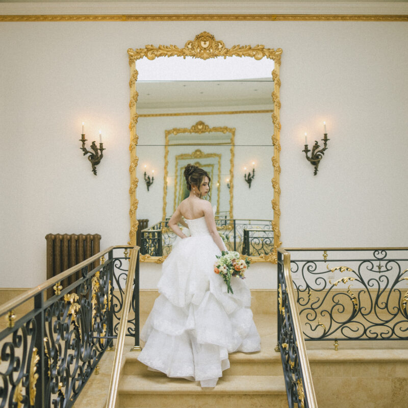 A bride in a strapless white gown stands on an elegant staircase during one of her bridal sessions, holding a bouquet. She is turned slightly, facing an ornate gold-framed mirror on the wall, with elegant sconces on either side, highlighting a classic, sophisticated setting.