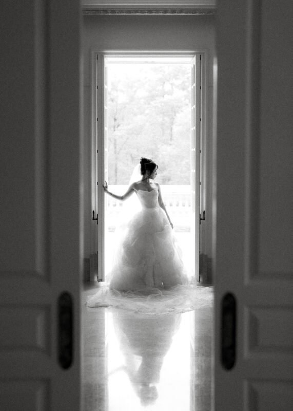 Black and white photo of a bride in a voluminous gown standing in a doorway, capturing the essence of timeless bridal sessions. Framed by open doors with light streaming in, a reflective pattern graces the polished floor. Outdoors, trees are visible in soft focus.