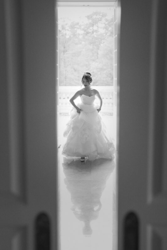 A bride during her bridal session stands at an open door, bathed in light. Her flowing gown is silhouetted against the bright outdoor backdrop, with soft shadows on the floor, crafting a serene and elegant scene.