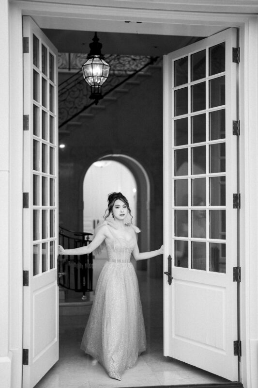 In an elegant strapless gown, a woman stands poised in a grand doorway during her bridal session. The black and white image captures the timeless beauty of the moment, with stairs and an ornate chandelier enhancing the sophisticated scene.