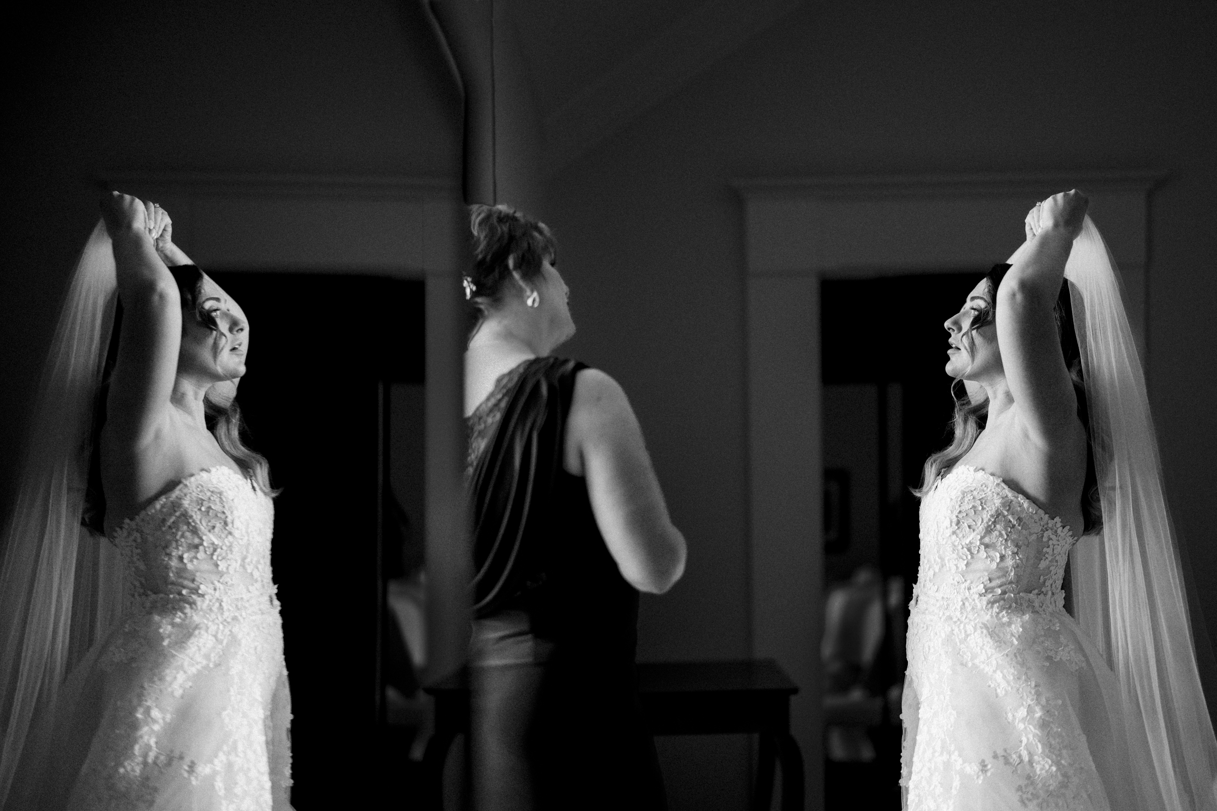 A bride in a wedding gown is adjusting her veil in front of a mirror, with the serene river landing just outside the window. A woman in a dark dress stands beside her, assisting. The room is dimly lit, creating a tranquil mood that perfectly complements the setting.