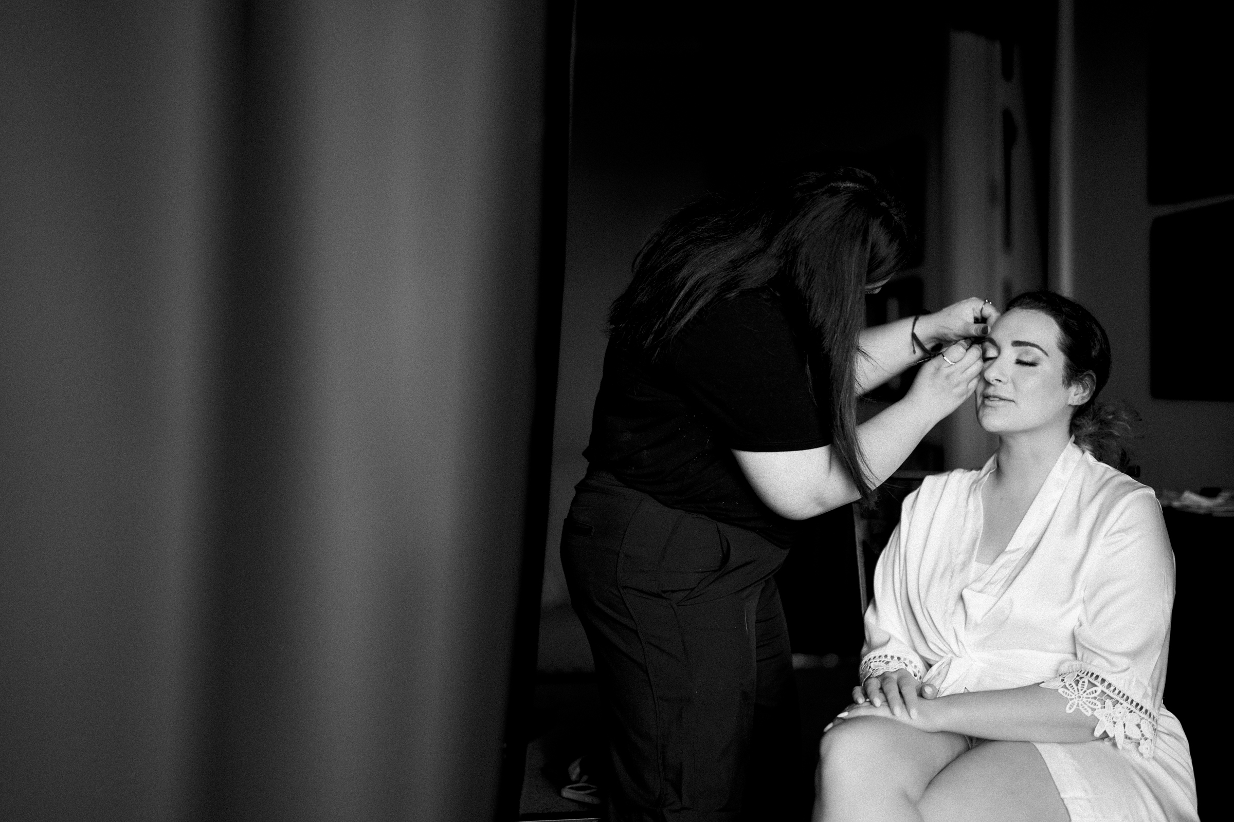 A woman sits with her eyes closed, wearing a robe, in the intimate and serene setting of River Landing. Another person stands beside her applying makeup, capturing the tranquil preparation for a beautiful wedding in timeless black and white.