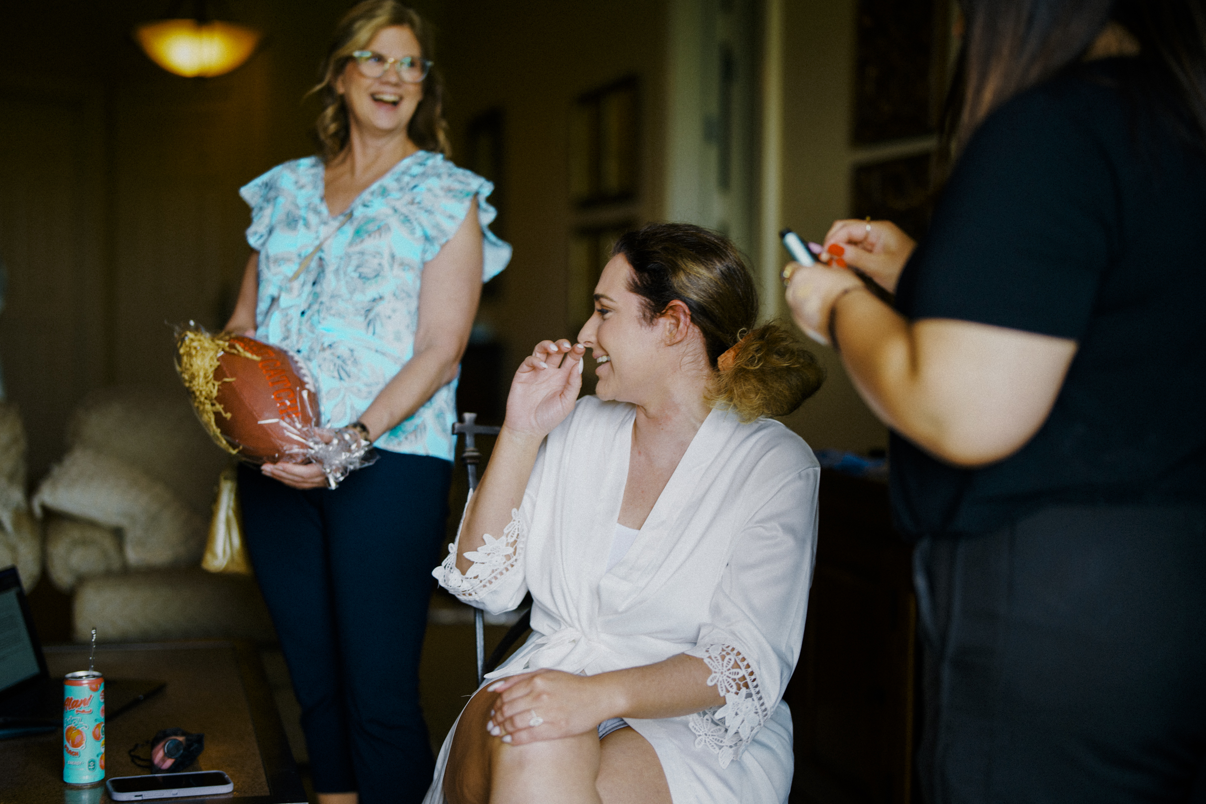 Three women in a room, one seated in a white robe laughing, another holding a football-shaped gift, and a third using a smartphone. A soft drink can is on the table nearby. The mood is relaxed and cheerful, reminiscent of an early gathering before a joyful river landing wedding.