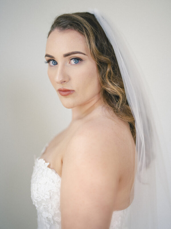 A bride with long, wavy hair and a white veil gazes over her shoulder at the river landing. She wears a strapless lace wedding dress and has a calm, focused expression. The softly blurred background emphasizes her serene and poised appearance.