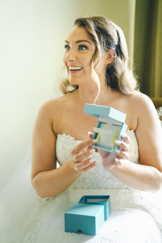 A bride in a white lace wedding gown smiles brightly, holding an open turquoise gift box that reveals a ring. She's sitting indoors at the River Landing wedding venue, with natural light beautifully illuminating her face.