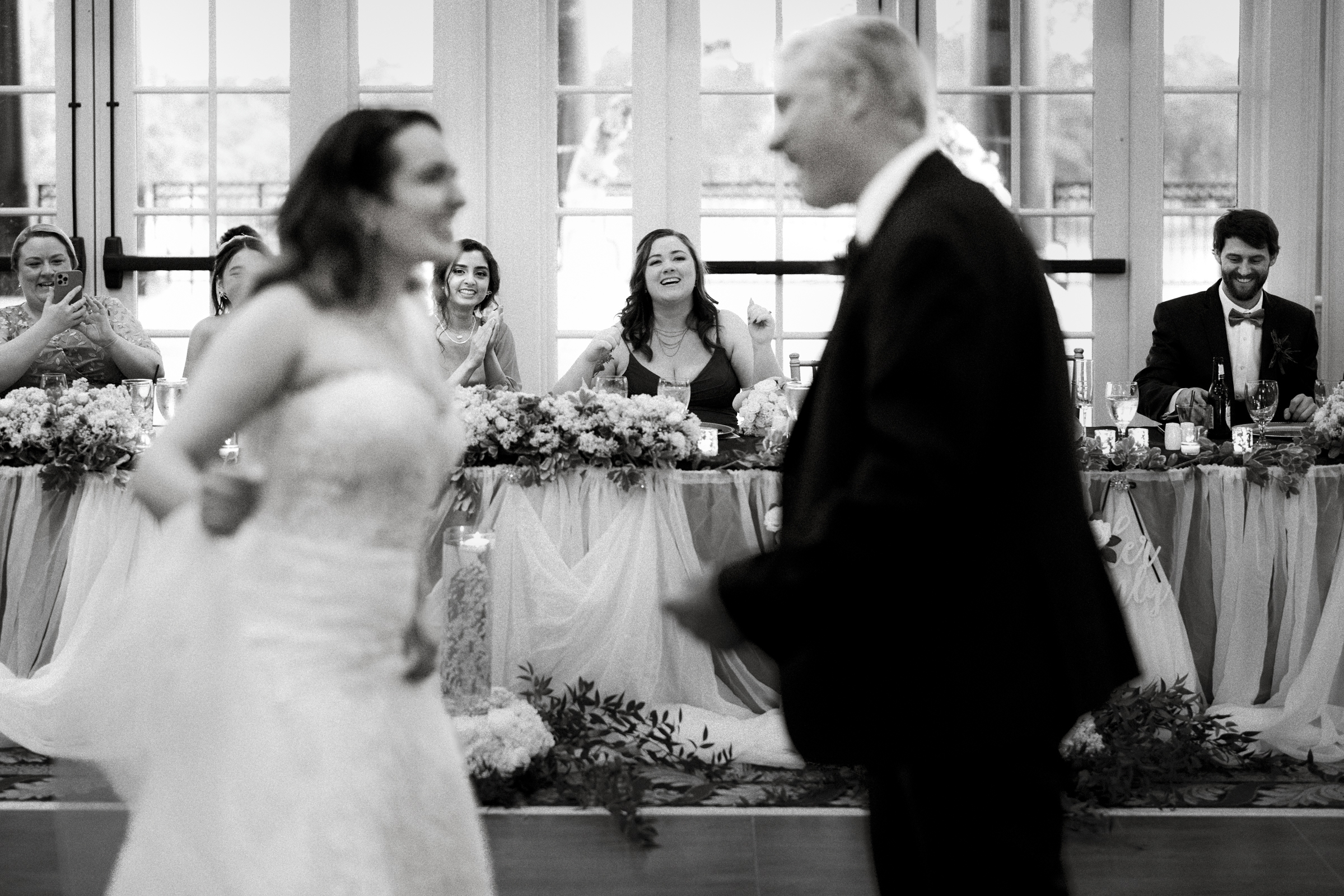 A bride and a man gracefully dance together in the enchanting River Landing wedding reception hall, as guests seated at a beautifully decorated table in the background cheer and smile. The black and white image captures a joyful moment.